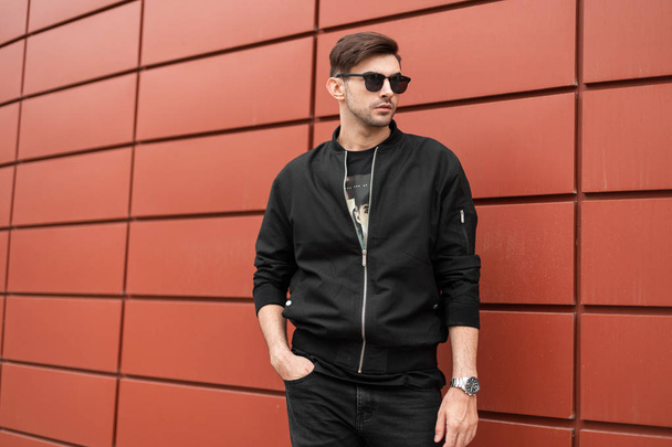 Moderno modelo de hombre joven y elegante en chaqueta negra de moda en gafas de sol vintage en jeans con un reloj de plata posando cerca de una pared de metal rojo en la ciudad. Un tipo americano atractivo. Estilo de calle
. - Foto, imagen
