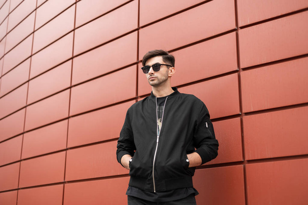 American attractive young hipster man with a fashionable hairstyle in stylish sunglasses in a vintage black jacket posing near a metallic red wall on the street. Handsome urban guy walks in the city. - Valokuva, kuva