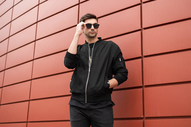 Attractive young hipster man in fashionable black clothes straightens trendy sunglasses. Stylish urban model guy stands outdoors in the city near the red metal wall. - Foto, imagen