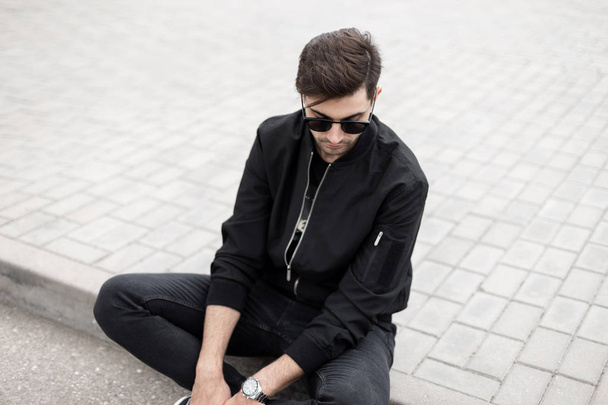 Attractive young hipster man in stylish sunglasses in black fashionable jacket in jeans is resting sitting on a tile on a street on a sunny summer day. Trendy american model guy relaxes outdoors. - Photo, Image