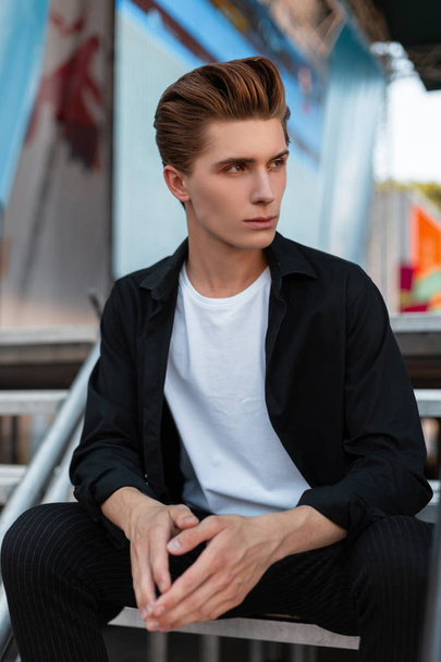 Ευρωπαίος όμορφος νέος άνδρας με ένα κομψό μαύρο πουκάμισο σε ένα λευκό T-shirt σε μοντέρνα παντελόνια με ένα κομψό χτένισμα που κάθονται σε μια μεταλλική σκάλα στην πόλη. Ελκυστικό τύπος μόδας μοντέλο. - Φωτογραφία, εικόνα