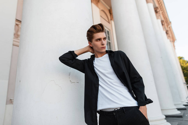 Vonzó fiatal férfi divatos frizura egy klasszikus fekete inget egy stílusos póló jelentő közelében egy épület szüreti oszlopokat. Jóképű modern fickó pihentető szabadban a városban. - Fotó, kép