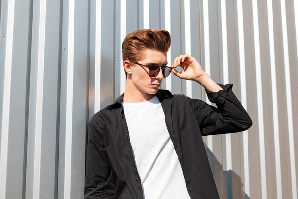 Houkutteleva nuori mies malli muodikas musta paita t-paita suoristaa tyylikäs aurinkolasit. Euroopan hipster kaveri poseeraa lähellä hopea metalli seinään kaupungissa valoisa aurinkoinen päivä. Moderni tyyli
 - Valokuva, kuva