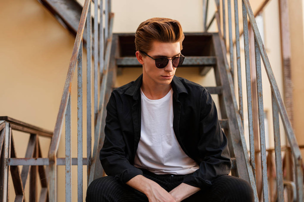 amerikanischer junger Hipster in elegantem Hemd und T-Shirt mit trendiger Sonnenbrille sitzt auf einer eisernen Treppe in der Stadt. schöner Kerl mit einer stylischen Frisur, die auf der Straße ruht. - Foto, Bild