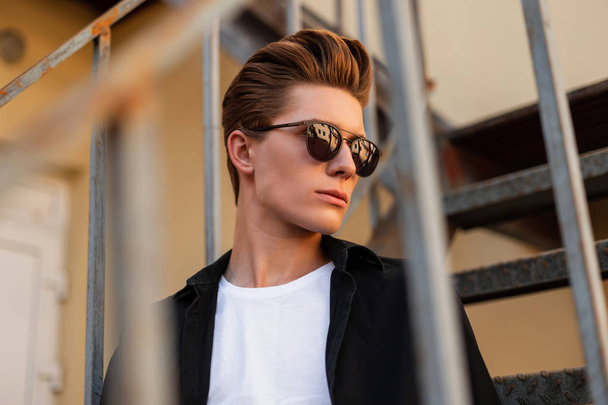 Portrait of a European serious young man with a fashionable hairstyle in stylish clothes in trendy sunglasses. Handsome guy hipster is sitting on a vintage iron staircase on the street at sunset. - Photo, Image