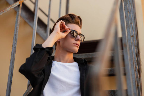 Young attractive hipster man straightens trendy black sunglasses. Portrait of a stylish handsome guy in a black vintage shirt with a fashionable hairstyle in the city on a summer day. European style. - Foto, Imagem
