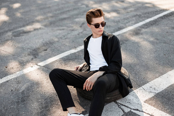Jeune homme hipster moderne en lunettes de soleil dans des vêtements à la mode avec une coiffure à la mode se détend assis sur la roue en caoutchouc de la voiture sur une rue dans la journée ensoleillée. Charmant mec qui se repose dans la rue
. - Photo, image