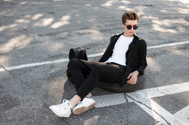 Handsome stylish hipster man relaxes sitting on the asphalt. Modern guy model in fashionable elegant black clothes in dark sunglasses with a black trendy backpack sits in the street on a sunny day. - Fotó, kép