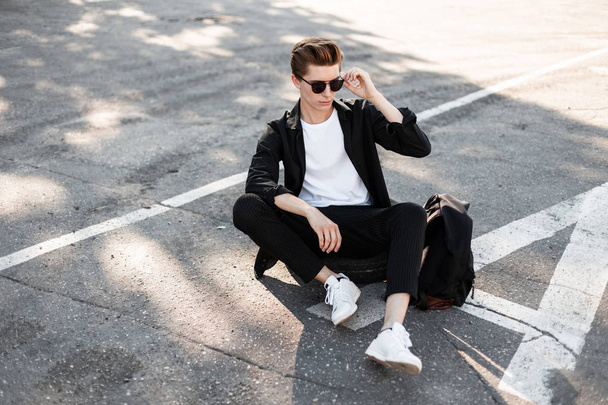 Hombre hipster joven urbano con peinado en gafas de sol en una camisa negra en pantalones a rayas de moda en zapatillas con una mochila sentarse en una rueda de goma en una calle en un día soleado. Un tipo guapo. Estilo de calle
. - Foto, imagen