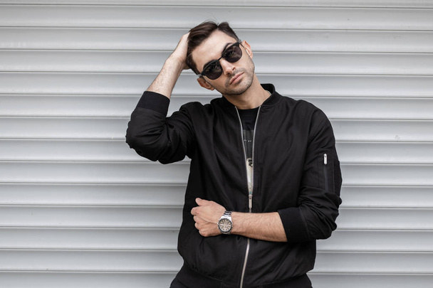 Handsome young hipster man in a fashionable black jacket in stylish sunglasses is posing outdoors near a white metal modern wall. Urban guy resting on the street. Street style. Trendy menswear. - Photo, Image