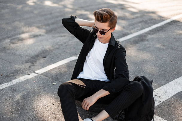 European young man with trendy hairstyle in elegant clothes in sunglasses with a backpack sits on a rubber wheel outdoors on a sunny day. Handsome guy hipster enjoys the summer sunshine. Street style. - Photo, Image