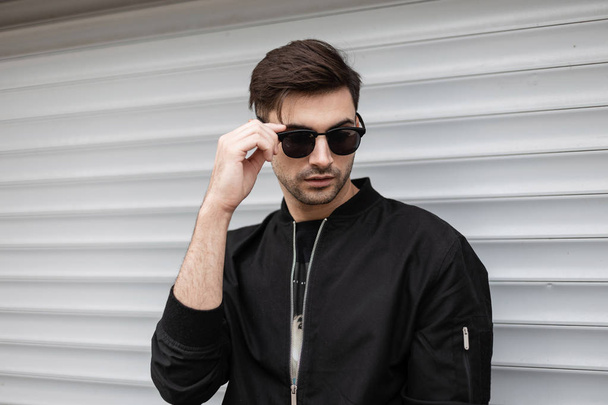 American hipster young man in a black stylish jacket stands and straightens trendy sunglasses near a metallic white wall on a summer day. Modern fashionable sexy guy brunette outdoors. - Zdjęcie, obraz