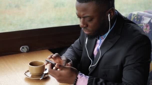 Czarny człowiek jest słuchanie muzyki w słuchawkach przeglądania smartphone siedzi w kawiarni. - Materiał filmowy, wideo