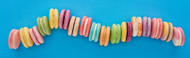 top view of multicolored delicious French macaroons in curved line on blue bright background, panoramic shot - Photo, Image