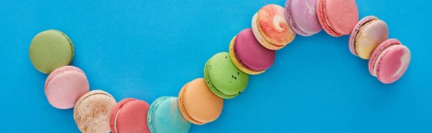 top view of multicolored delicious French macaroons in curved line on blue bright background with copy space, panoramic shot - Foto, imagen