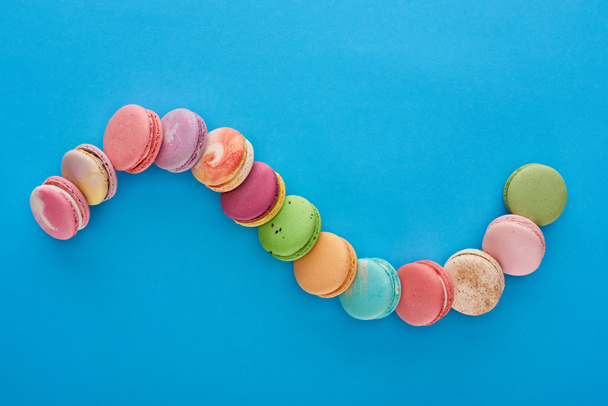 top view of multicolored delicious French macaroons in curved line on blue bright background with copy space - Photo, Image