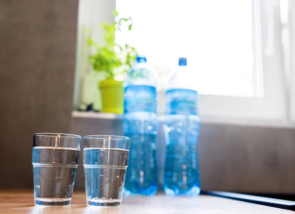 Two glasses of water in kitchen, with tap water and two bottles of mineral water. Shallow depth of field. Water shortage concept, saving water. Body hydration with clean & clear water. - Photo, Image