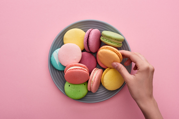cropped view of female hand near plate with multicolored delicious French macaroons on pink background - Foto, afbeelding