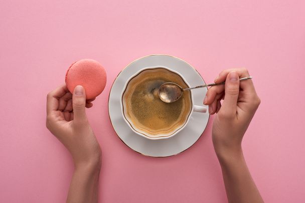 partial view of woman holding spoon near coffee in cup and delicious French macaroon on pink background - Fotó, kép