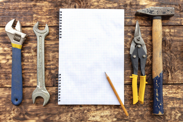 Un cuaderno en blanco con un lápiz rodeado de herramientas de mano sobre una vieja superficie de madera
. - Foto, imagen