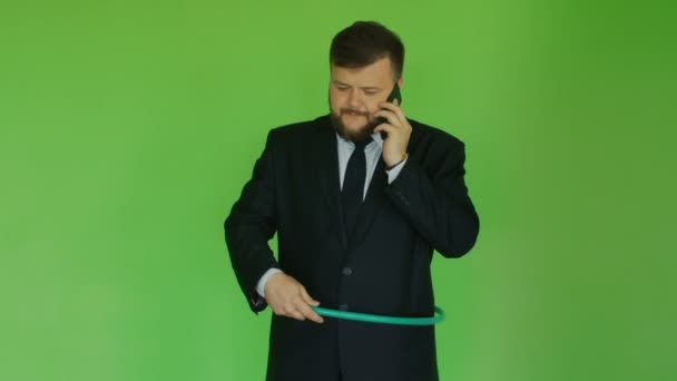 Plump businessman talking on the phone and trying to twist the hula hoop. Funny situation. Chromakey Green. Bearded thick guy in black jacket, shirt and tie solves problems by smartphone. Man - Séquence, vidéo