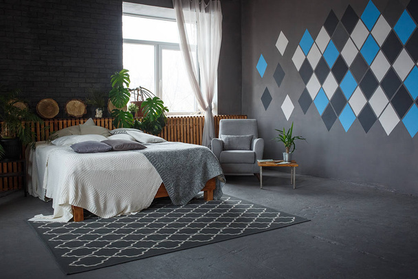 Стильна лофт затишна вітальня з двоспальним ліжком, килимом, кріслом, зеленими рослинами та геометричними візерунками на стіні
 - Фото, зображення