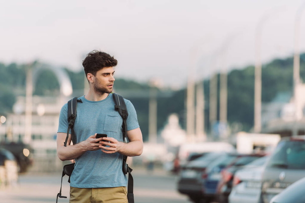 handsome man in t-shirt holding smartphone and looking away  - Fotó, kép