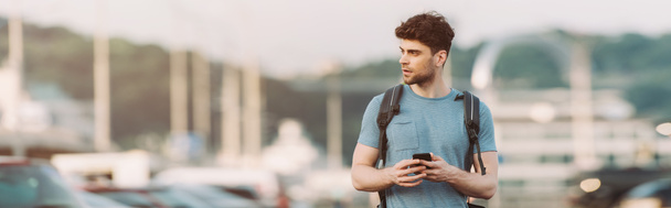 panoramic shot of handsome man in t-shirt holding smartphone and looking away  - Fotó, kép