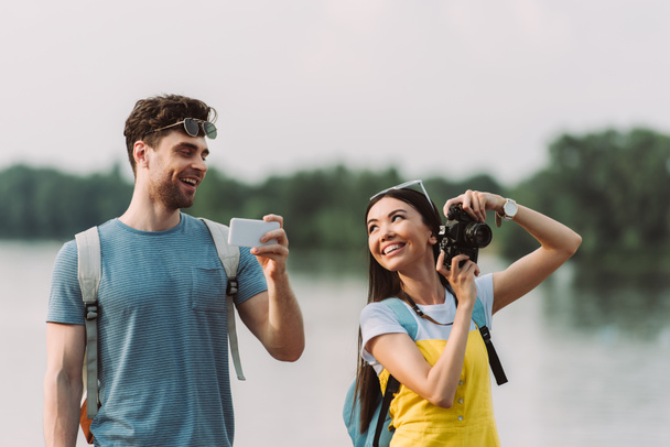smiling and asian woman holding digital camera and handsome man holding smartphone  - Fotó, kép
