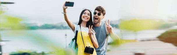 panoramic shot of handsome man and asian woman showing peace signs and taking selfie  - Valokuva, kuva