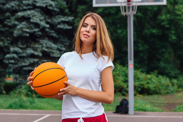 Beautiful young girl dressed in white t-shirt, shorts and sneakers, plays with a ball on a basketball court. - Photo, image