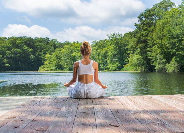 Attractive woman is practicing yoga and meditation, sitting in lotus pose near lake in morning. - Photo, Image