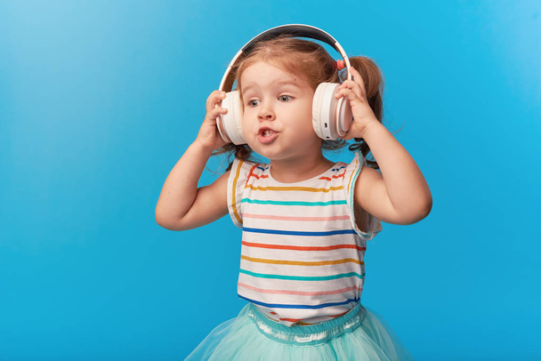 Happy smiling child enjoys listens to music in headphones over colorful bleu background. Vivid and fun emotions, happy child with pleasure listens to songs in wireless headphones - Foto, afbeelding