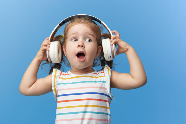 幸せな笑顔の子供は、カラフルなブルーの背景の上にヘッドフォンで音楽を聴いて楽しんでいます。鮮やかで楽しい感情、喜びを持つ幸せな子供は、ワイヤレスヘッドフォンで曲を聴きます - 写真・画像
