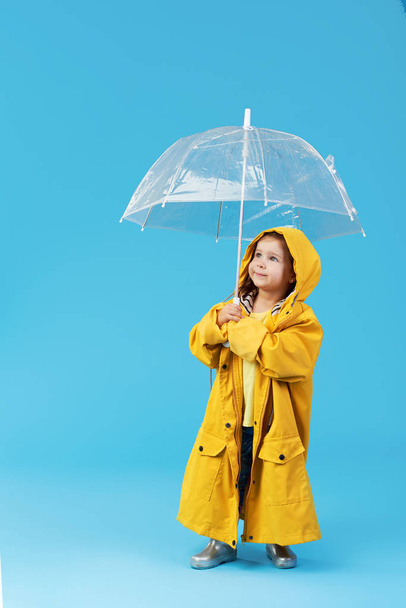 Gelukkig grappig kind met transparante paraplu poseren op blauwe studio achtergrond. Het meisje draagt een gele regenjas en rubberen laarzen. Heeft een vintage reiskoffer - Foto, afbeelding