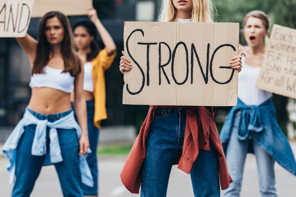 обрезанный вид феминистки, держащей плакат с надписью сильная рядом с женщинами на улице
 - Фото, изображение