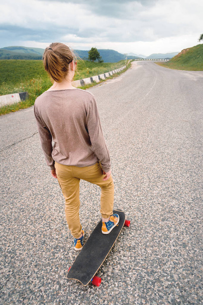 Young stylish man with long hair gathered in a ponytail and in sunglasses stands with a longboard on a country asphalt road in the mountains on the background of epic rocks - Photo, image