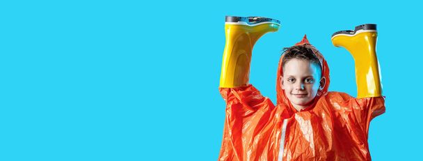 smiling boy in orange raincoat stuck his hands in rubber boots on blue background - Photo, Image