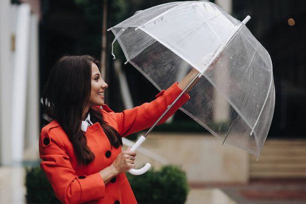 Woman opening closing umbrella. Portrait of a long-haired stylish young smiling girl waiting for rain in red coat looking at the sky on city background. - Fotografie, Obrázek