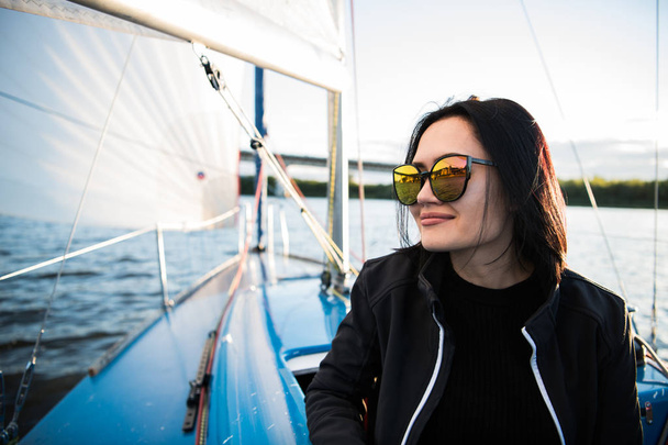 Gyönyörű fiatal nő ül a jacht tálban, és pózok. Visel sunglusses kézzel és mosolyogva. Modell vitorlás fedélzetén Yacht. - Fotó, kép