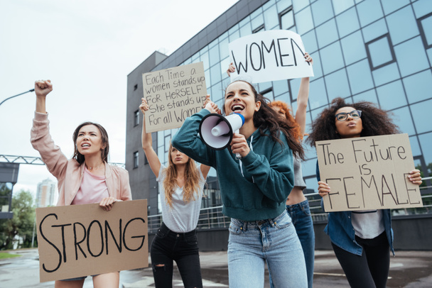 selective focus of emotional girl holding megaphone and screaming near multicultural women holding placards on meeting  - Photo, Image
