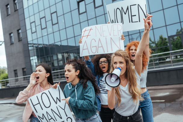 enfoque selectivo de chica emocional sosteniendo el megáfono y el gesto cerca de las mujeres multiculturales con pancartas en la reunión
  - Foto, Imagen