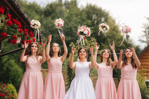 Happy bride with bridesmaid hold bouquets and have fun outside. Beautiful bridesmaid in same dresses stand by the charming bride in long wedding dress - Photo, Image