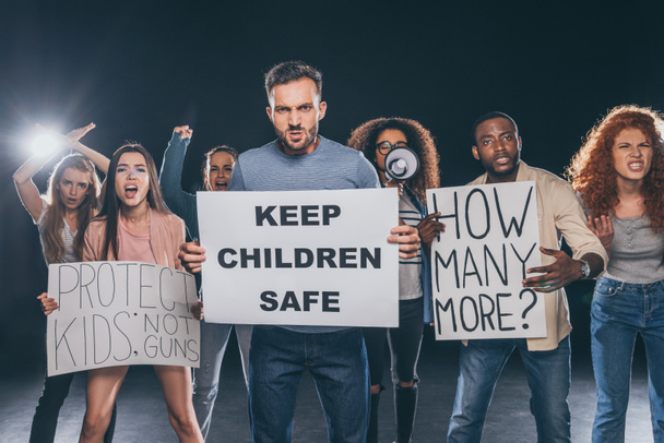 hombre enojado sosteniendo pancarta con mantener a los niños letras seguras cerca de personas multiculturales en negro
  - Foto, Imagen