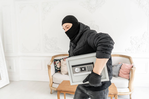 The burglar commits a crime in Luxury apartment with stucco. Thief with black balaclava stealing modern Electronic safe box. - Fotoğraf, Görsel