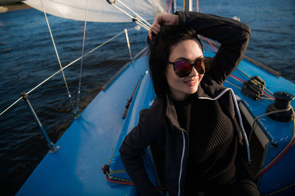 Mooie jonge vrouw zit op Yacht Bowl en poses. Ze draagt sunglusses met de hand en glimlach. Model is zeilen aan boord van Yacht. - Foto, afbeelding