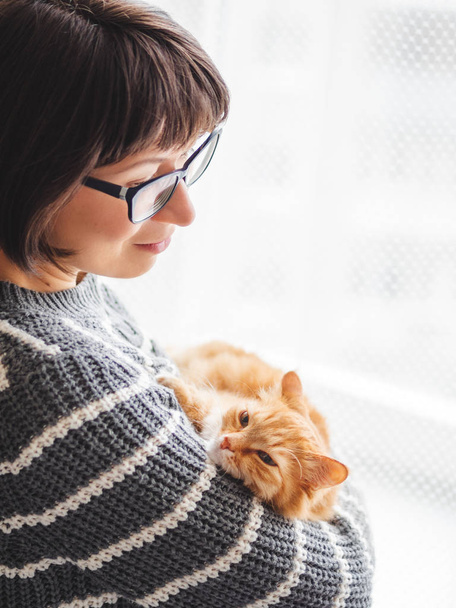 Cute ginger cat dozing on woman arms. Smiling woman in grey knitted sweater stroking her fluffy pet. Cozy home. - Фото, изображение