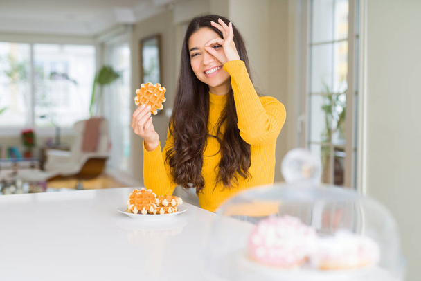 Jeune femme mangeant de la pâte à gaufres sucrée avec le visage heureux souriant faire ok signe avec la main sur les yeux en regardant à travers les doigts
 - Photo, image
