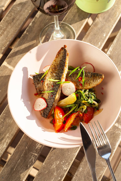 gebratener Makrelenfisch mit Gemüsegarnitur: Spinat in Kokosmilch, Rettich, Zwiebeln und gebackenen Tomaten, serviert auf einer sonnigen Terrasse mit einem Glas Rotwein - Foto, Bild