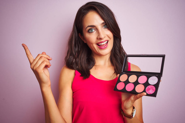 Young beautiful woman applying make up over pink isolated background very happy pointing with hand and finger to the side - Фото, изображение